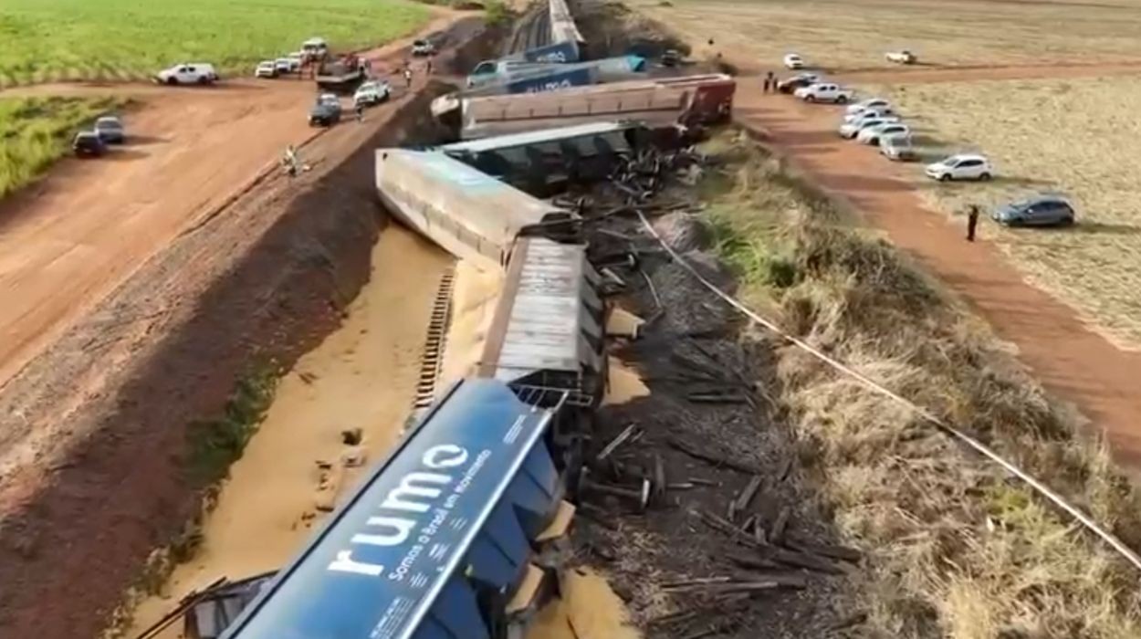 Vagões de trem de carga descarrilados e tombados ao lado da linha férrea, com milho espalhado no chão, após colisão com caminhão.