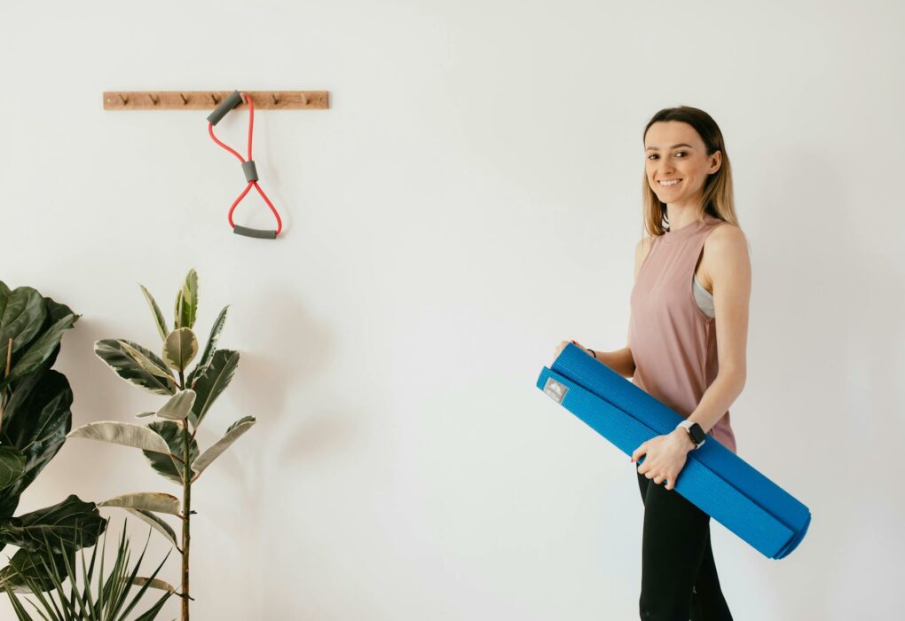 Smiling woman holding a blue yoga mat, preparing for a fitness session at home, emphasizing a healthy lifestyle.