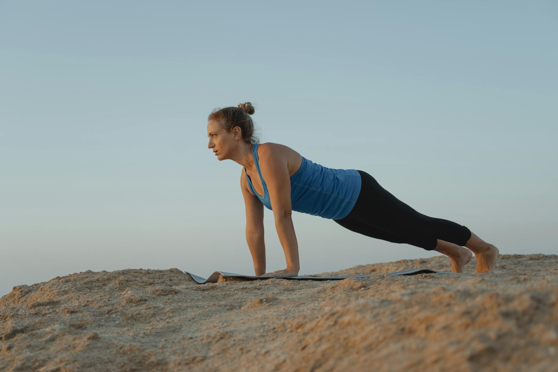 A woman performs a yoga pose on a sandy outdoor terrain under a clear sky at dusk.