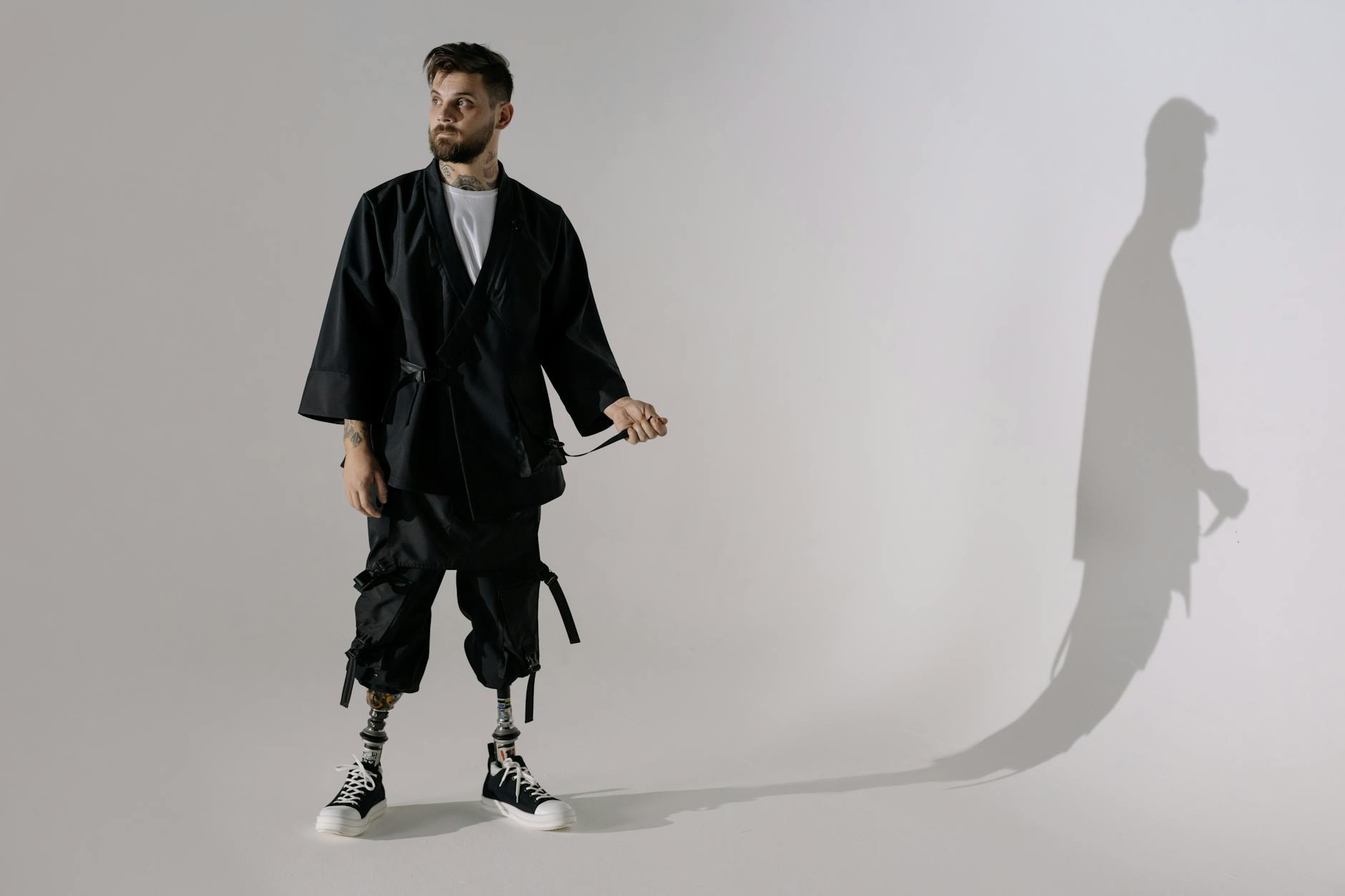 A modern, stylish man with prosthetic legs wears black clothing and sneakers in a studio setting.