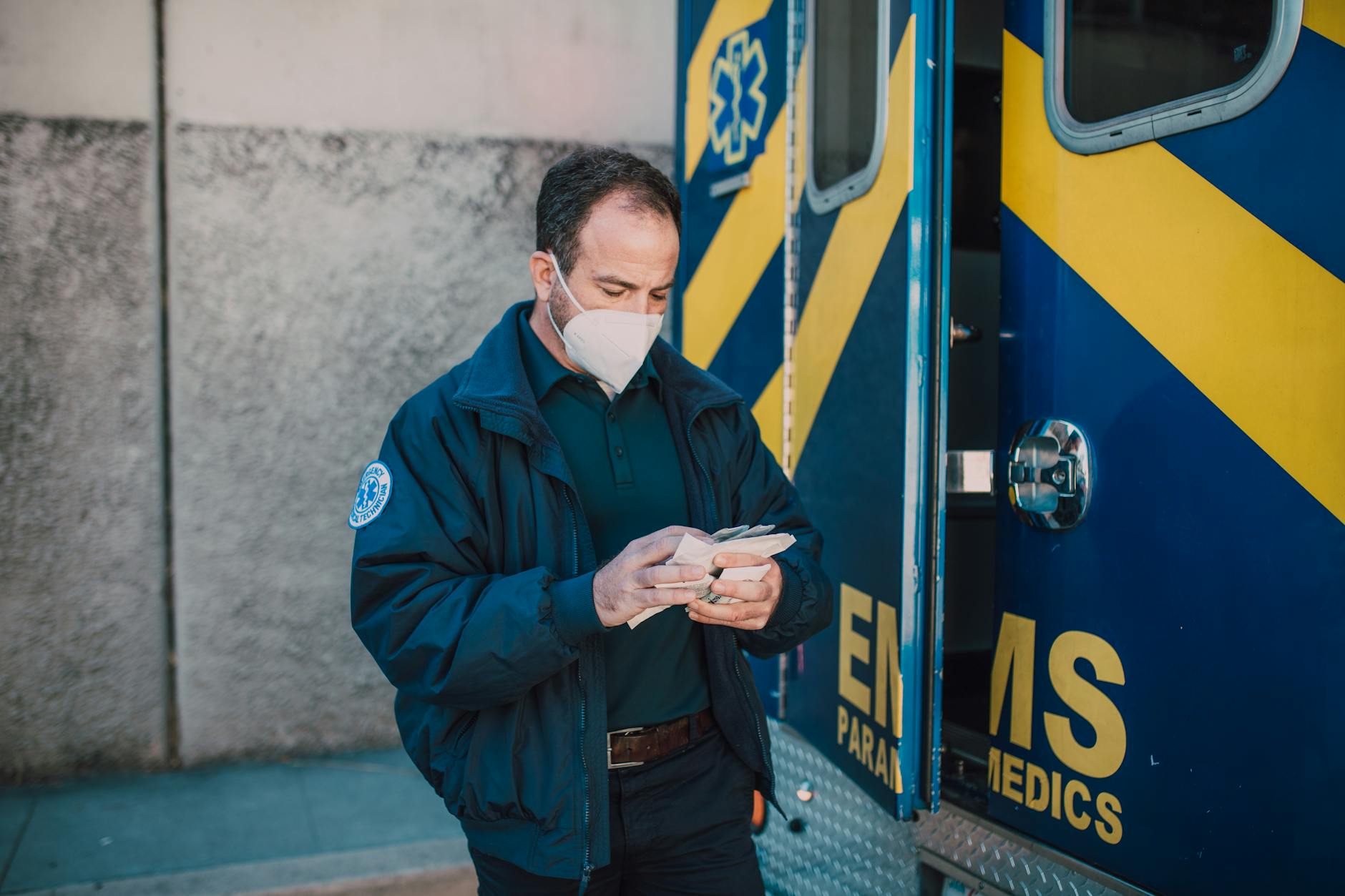 Paramedic in mask working next to EMS vehicle, preparing for emergency response.
