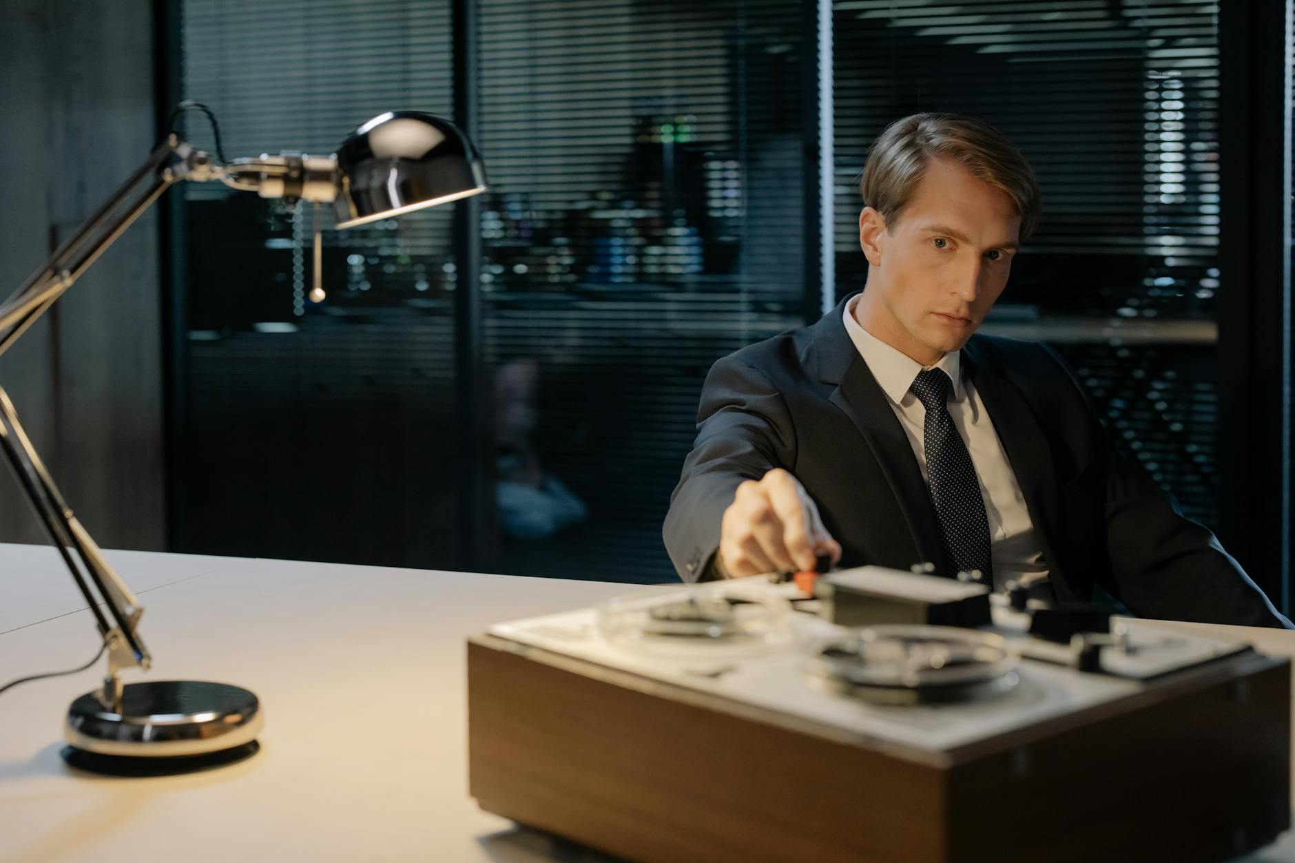 A detective in a suit uses a tape recorder under a desk lamp, evoking vintage investigation themes.